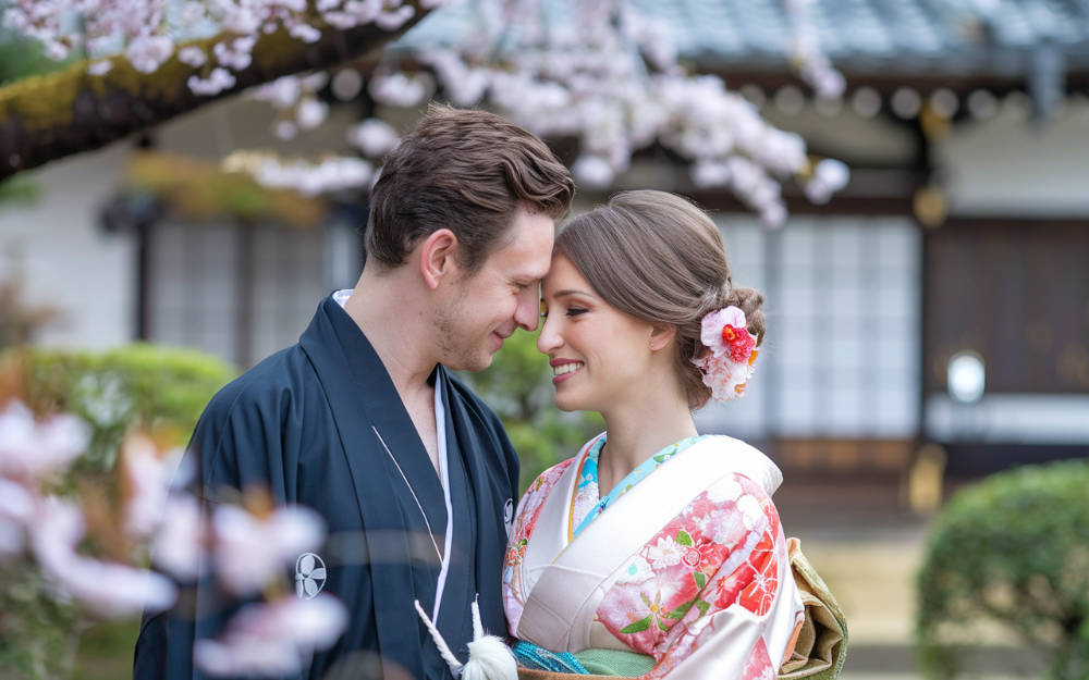 A high-quality close-up shot of a couple enjoying their luxury honeymoon in Japan. The man is wearing a traditional Japanese robe, and the woman is wearing a kimono. They are standing in a garden with cherry blossoms. The background contains a traditional Japanese building. The image has a soft focus.