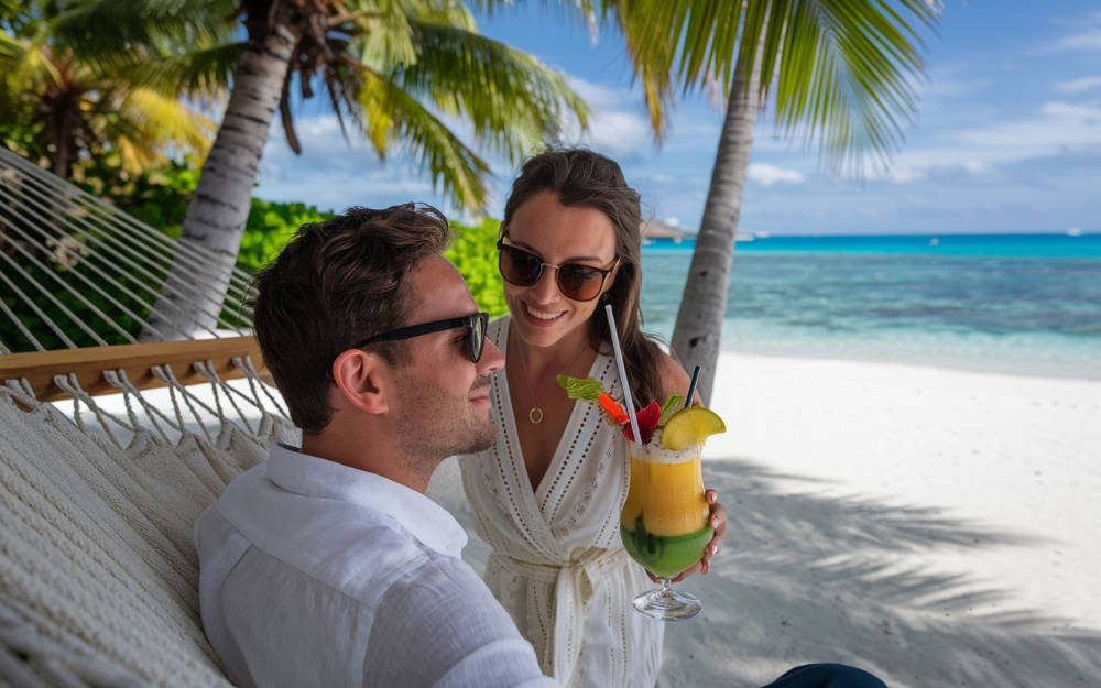 A high-quality close-up shot of a couple enjoying their luxury honeymoon in the Seychelles. The man is sitting in a hammock between two palm trees, with the ocean in the background. He is wearing a white shirt and sunglasses. The woman is standing near him, wearing a white dress and sunglasses. She is holding a tropical drink with a straw. The background is the pristine white sand beach and the clear turquoise water of the Seychelles.